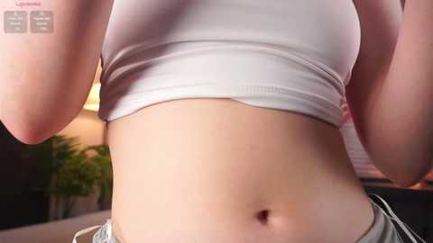 Media: Video of a fair-skinned woman with a flat stomach, wearing a white crop top and gray pants, standing indoors with blurred background featuring a potted plant.