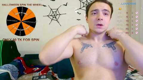 Media: Video of a shirtless, fit, young man with a surprised expression, wearing tattoos of two pistols on his chest, standing in a messy room with Halloween decorations, including a dartboard, spiderweb, and bats.