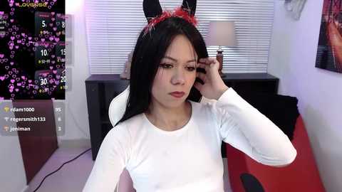 Media: Video of a young woman with long black hair, wearing a white long-sleeved shirt and bunny ears headband, adjusting her hair in a modern, cozy room with a lamp, purple background, and social media overlay.