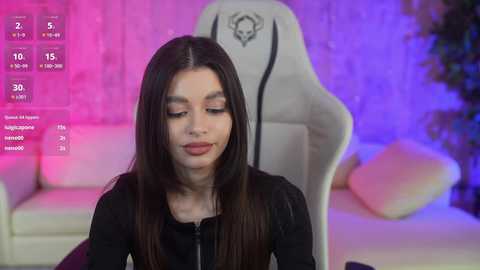 Media: Video of a young woman with long brown hair, wearing a black top, seated in a white gaming chair with a skull logo, in a room with purple and pink lighting.