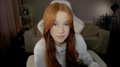 Media: Video of a young red-haired girl in a white gamer chair with a \"F\" logo, wearing a white long-sleeve shirt, indoors, dimly lit room with beige couch, shelves, and plants.