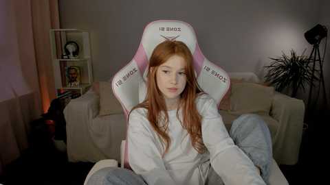 Media: Video of a young Caucasian woman with long, straight red hair, sitting in a pink and white gaming chair, wearing a white hoodie, in a dimly lit living room with a bookshelf and plants.