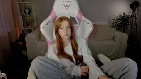 Media: Video of a young red-haired girl in a white and pink gaming chair, wearing a white hoodie and gray sweatpants, in a cozy living room with a beige couch and bookshelves.