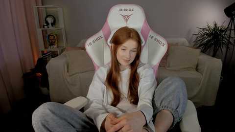 Media: Video of a young, fair-skinned redhead with long hair, wearing a white sweatshirt and grey sweatpants, seated in a pink and white gaming chair with \"Twitch\" branding, in a cozy, dimly lit living room.