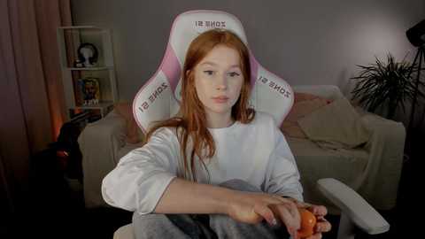 Media: Video of a young, pale-skinned woman with long red hair, seated in a pink gaming chair, holding a controller, in a dimly lit, cozy living room with a beige sofa, shelves, and plants.