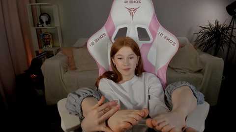 Media: Video of a young red-haired girl with pale skin sitting in a pink gaming chair, wearing a white shirt and gray shorts, in a cozy living room with a bookshelf and a cat.