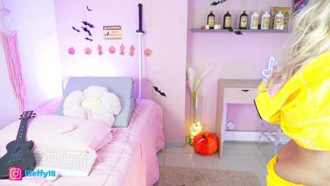 Media: Video of a young blonde woman in a yellow crop top, standing in a pastel-themed room with a pink sofa, a guitar, and Halloween decorations.