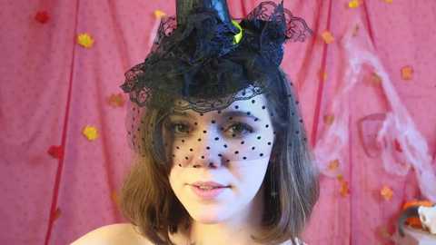 Media: Video of a young woman with shoulder-length brown hair wearing a black lace veil with polka dots, a black feathered headpiece, and a pink floral backdrop.