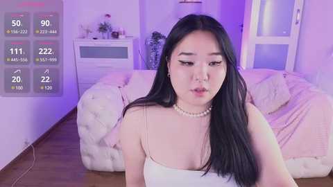 Media: Video of an East Asian woman with long black hair, wearing a white spaghetti-strap dress and pearl necklace, sitting in a plush, pink couch-filled living room with purple lighting.