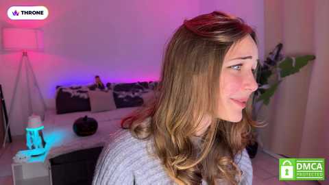 Media: Video of a young woman with long, wavy brown hair, smiling, in a modern living room with purple and blue lighting, a dark sofa, and a lamp.