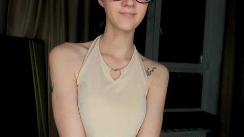 Media: Video of a slender, fair-skinned woman with glasses, wearing a beige halter top, revealing a small heart-shaped cutout on the chest. She has a tattoo on her right shoulder. Background shows a dark room with a window and curtains.