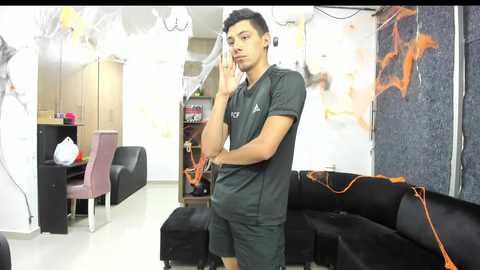 Media: Video of a young man with short black hair, wearing a gray T-shirt and shorts, standing in a modern, minimalist living room with black couches, white walls, and abstract art.