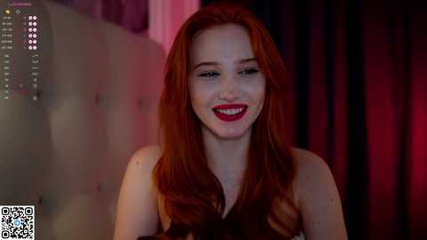 Media: Video of a smiling woman with long red hair, wearing red lipstick, in a dimly lit room with a tufted headboard.