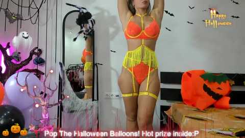 Media: Video of a woman in neon orange lingerie with yellow fringe, posing in a Halloween-themed bedroom with a pumpkin, spider web decor, and \"Happy Halloween\" text.