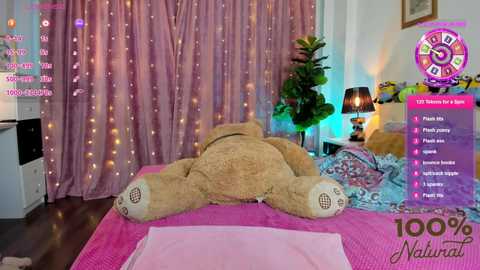 Media: Video of a cozy bedroom with a large, fluffy teddy bear on a pink bedspread, surrounded by string lights, a green plant, and a purple clock.