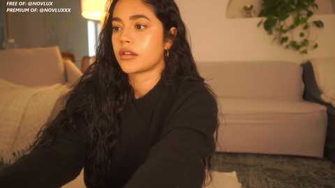 Media: Video of a young woman with long, wavy black hair and medium skin tone, wearing a black sweater, sitting on a beige couch in a cozy living room.