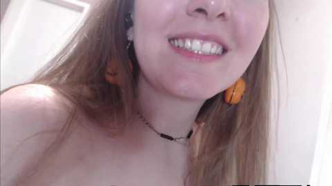 Media: Video of a young woman with fair skin, long brown hair, and a big smile, wearing orange earbuds and a black necklace. She is topless, with her bare shoulders visible.