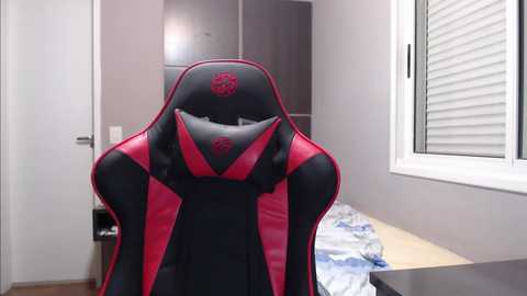 Media: Video of a black and red gaming chair with a high backrest in a minimalist bedroom, featuring a white window with blinds, a white bed with blue sheets, and a wooden floor.