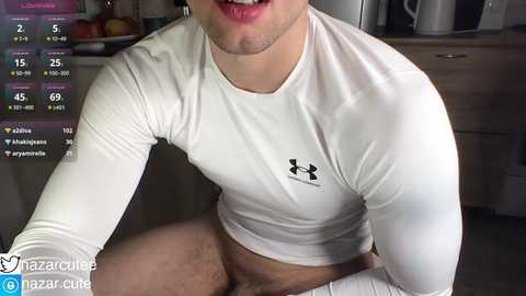 Media: Video of a fit, light-skinned man in a white Under Armour shirt, sitting in a kitchen with a wooden counter and fruit bowl, displaying his muscular physique and slightly open mouth.