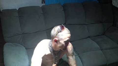 Media: Video of a shirtless, muscular, tattooed man with a bandana and sunglasses, sitting on a beige couch in a dimly lit room.