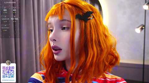 Media: A video of a young Asian woman with bright orange hair, wearing a striped shirt, and a black hair clip. She appears in a modern, dimly lit room with a digital screen displaying information.