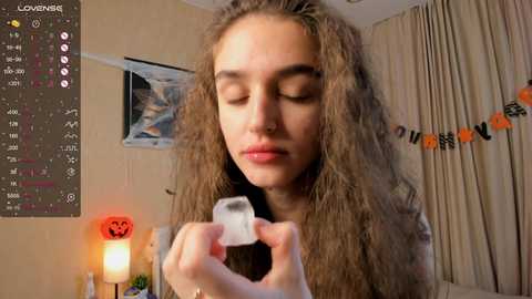 Media: Video of a young woman with long, wavy brown hair, eyes closed, holding a clear crystal in her hands, in a cozy room with Halloween decorations, including a pumpkin candle and orange garland.