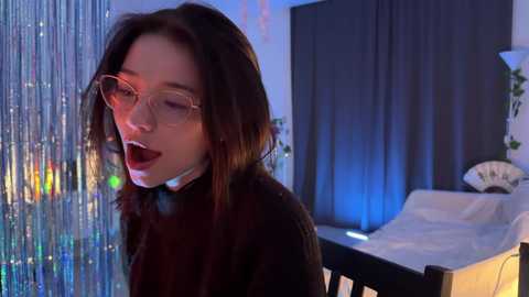 Media: A video of a young woman with a fair complexion, long brown hair, and clear glasses, wearing a black sweater, in a dimly lit room with blue and red lights, and a bed with white sheets in the background.