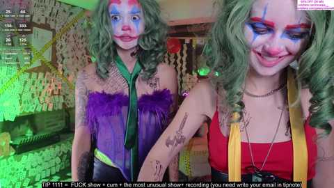 Media: Video of a woman with green hair, wearing a colorful clown costume, posing with a playful expression in a dimly lit room with graffiti and a green light.