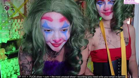 Media: Video of two women in cosplay, one with green hair and blue face paint, the other with red face paint and green hair. Both wear revealing outfits. Background is colorful and chaotic.