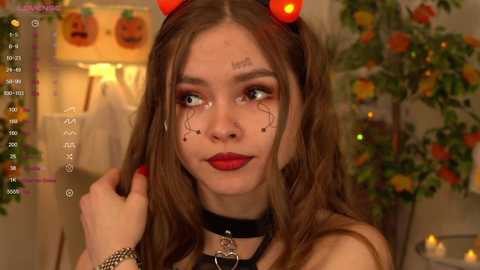 Media: Video of a young woman with fair skin and brown hair, wearing a black choker and devil horns, with Halloween-themed makeup, in a cozy, Halloween-decorated room.