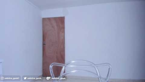 Media: A minimalist, white-walled room with a wooden door, a clear acrylic chair, and a small, white table. The room is sparsely furnished with a modern, clean aesthetic.