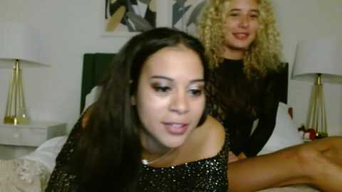 Media: Video of a young woman with long dark hair in a black glittery dress, seated on a bed, smiling; another woman with curly blonde hair in a black top stands behind her, laughing.