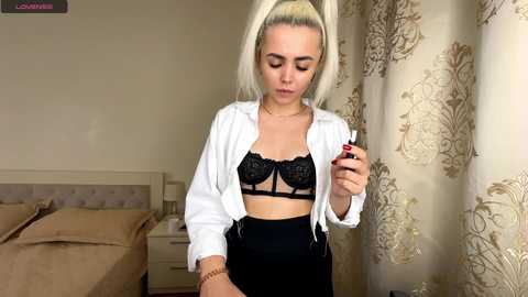 Media: Video of a blonde woman in black lace bra and high-waisted pants, holding a cigarette, standing in a beige bedroom with floral curtains.