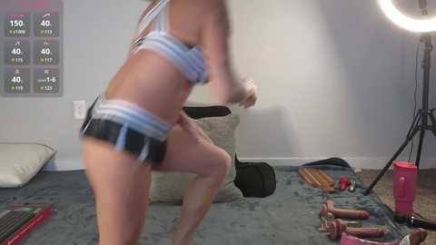 Media: Video of a fit woman in a white sports bra and black shorts, mid-jump, in a gym with weights, equipment, and a motivational poster in the background.