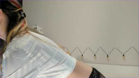 Media: Video of a person with light skin and long, dark hair partially tied back, wearing a white shirt and black shorts, standing against a plain white wall with a string of Edison-style light bulbs.