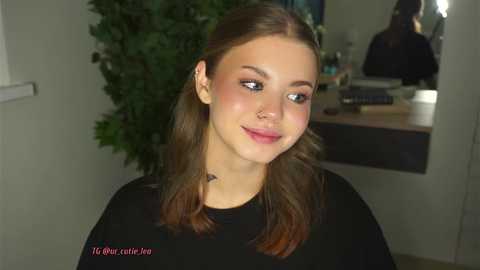 Media: Video of a young Caucasian woman with fair skin, brown hair, and freckles, wearing a black top. She has a subtle makeup look with pink blush. Background includes a potted plant and a mirror.