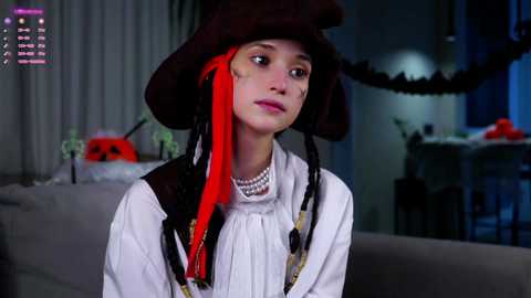 Media: Video of a young woman with pale skin and red hair, dressed in a historical costume with a black hat and white blouse, in a dimly lit room with Halloween decorations.