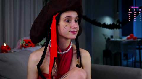 Media: Video of a young woman with pale skin, wearing a pirate hat, red dress, and pearl necklace, seated indoors. Background includes festive decor, candles, and a table.