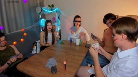 Media: Video of six people, three men and three women, playing poker at a wooden table in a dimly-lit room with neon lights, wearing casual attire and holding cards.