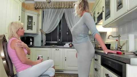 Media: Video of a blonde woman in a pink top and white pants sitting at a kitchen table, while a blonde woman in a light blue sweater and gray pants stands at the stove, both in a modern kitchen.