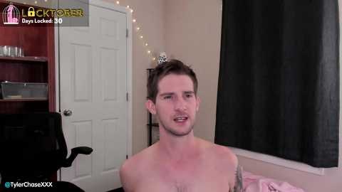 Media: Video of a young, shirtless man with short brown hair and a tattoo on his chest, standing in a bedroom with beige walls, a closed door, and a black curtain.