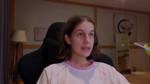 Media: Video of a young girl with long brown hair, wearing a white shirt, sitting in a black gaming chair in a dimly lit room with a lamp, shelves, and a circular mirror.