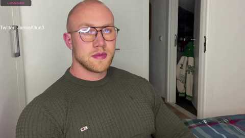 Media: Video of a bald, muscular white man with glasses, wearing a green, long-sleeve shirt, standing in a bedroom with white doors and a partially open closet showing laundry.