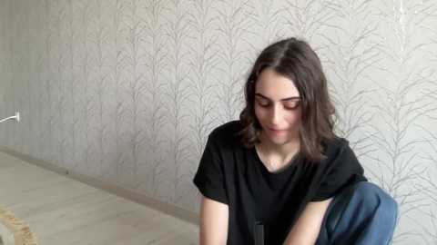 Media: Video of a young woman with shoulder-length brown hair, wearing a black t-shirt, sitting on a blue chair in a room with light gray patterned wallpaper.