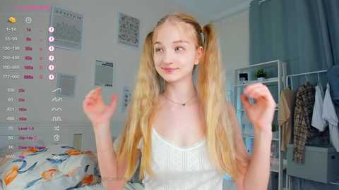 Media: A video of a young, fair-skinned girl with long blonde pigtails, wearing a white tank top, smiling and playfully posing in a bright, tidy bedroom with a bed and shelves.