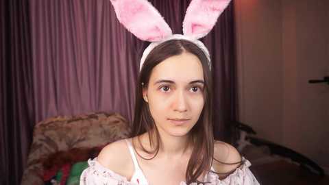 Media: Video of a young woman with light skin and long brown hair, wearing a fluffy pink bunny headband and off-shoulder white lace dress, standing in a dimly lit, cozy room with purple curtains and a floral-patterned couch in the background.