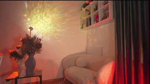 Media: Video of a cozy living room with a white sofa, a decorative vase of dried flowers on a black table, a glowing yellow light installation on the wall, and red curtains.