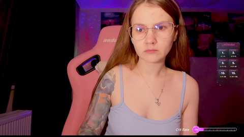 Media: A young woman with pale skin, long brown hair, and glasses sits in a pink gaming chair, wearing a light blue tank top. She has colorful tattoos on her left arm. The room is dimly lit with purple and blue hues, featuring gaming posters on the wall.