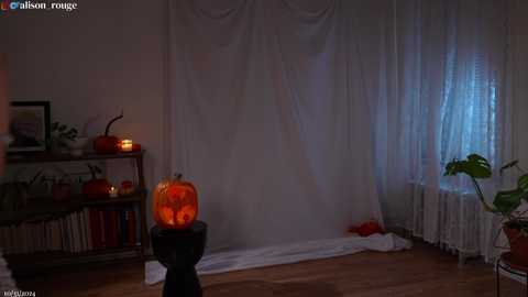 Media: A dimly lit, Halloween-themed room with a carved pumpkin on a pedestal, a lit jack-o'-lantern, white curtains, and a small potted plant.