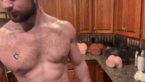 Media: Video of a shirtless, muscular, light-skinned man with a beard, tattoo, and a toned physique, standing in a kitchen with wooden cabinets, granite countertops, and kitchenware, including a coffee maker and bread rolls.